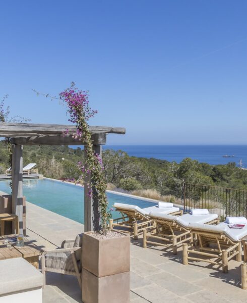 A scenic outdoor pool area with cushioned lounge chairs overlooks a vast, clear blue ocean, epitomizing luxury travel. The pool is bordered by a wooden pergola adorned with flowering vines, while lush greenery surrounds the area under a clear blue sky. David Pearson Travel