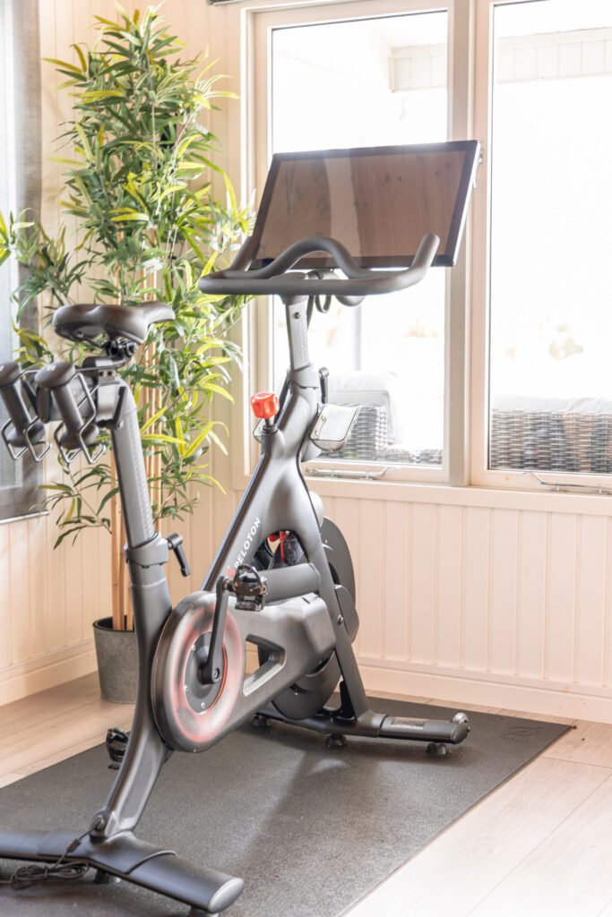 A stationary Peloton exercise bike with a large touchscreen is set up indoors on a black mat near a window, next to a tall potted plant. Sunlight is streaming in from outside. David Pearson Travel