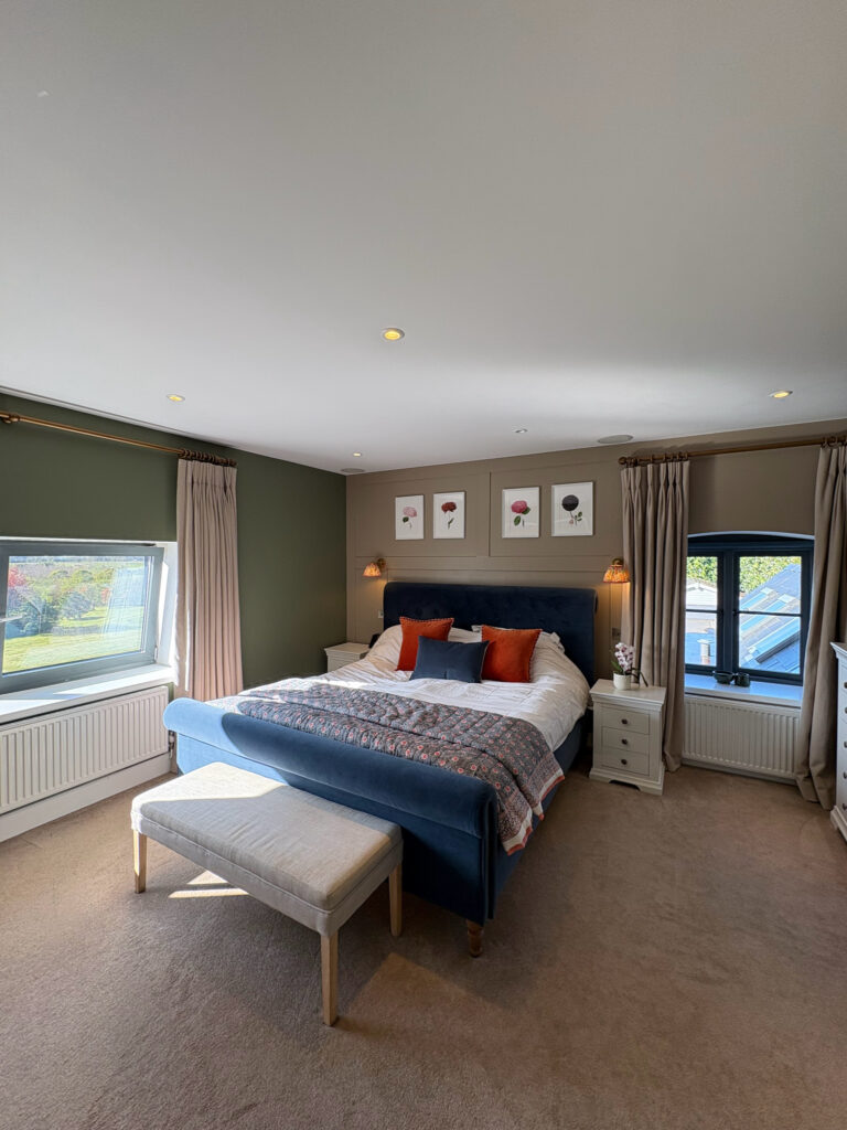 A bright, modern bedroom with a blue bed, white linens, and orange pillows. Three flower prints hang above the headboard. Two windows with beige curtains let in natural light. A white bench sits at the foot of the bed. David Pearson Travel