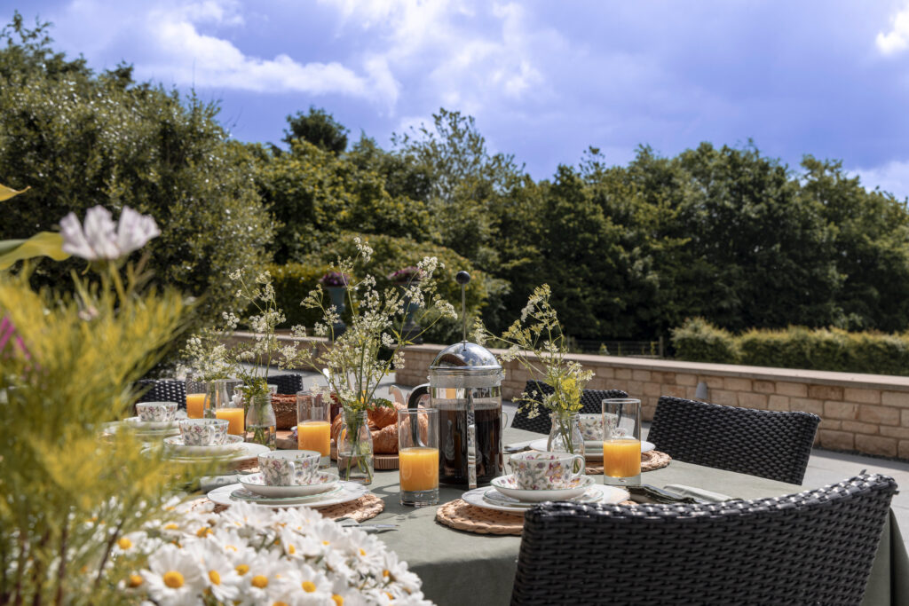 An outdoor table set for breakfast with floral dishes, glasses of orange juice, a French press, and fresh flowers. The table is surrounded by wicker chairs and greenery, under a partly cloudy sky. David Pearson Travel