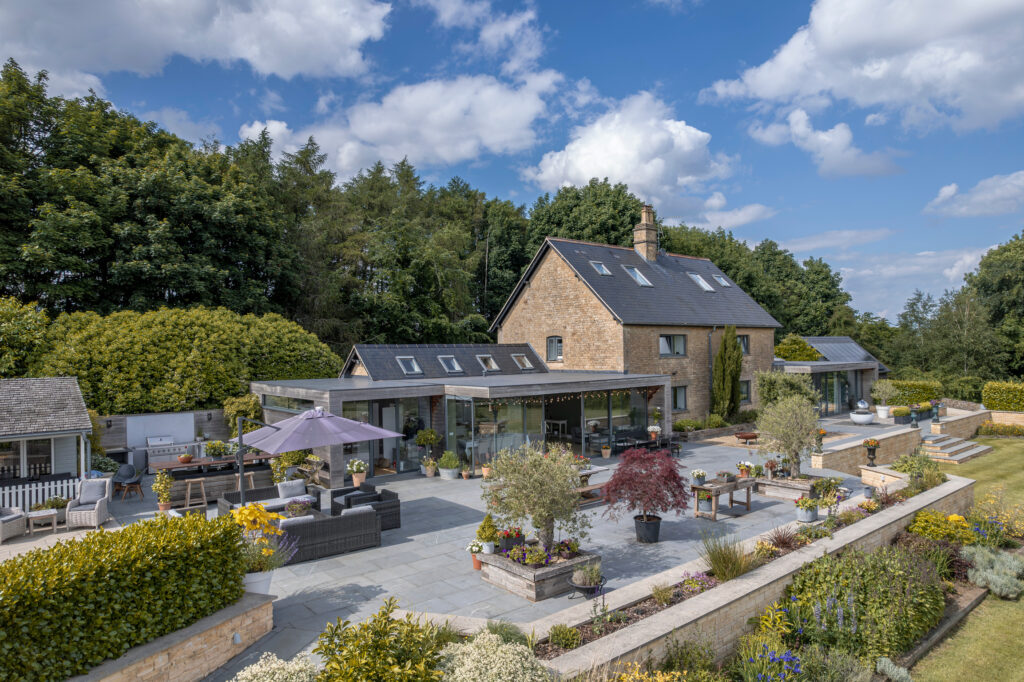 A modern stone house with a large patio, outdoor seating, and landscaped gardens, surrounded by trees and greenery under a blue sky with scattered clouds. David Pearson Travel