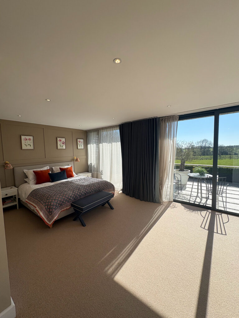 Bright bedroom with a large bed, colorful pillows, and framed art above the headboard. Sunlight streams in through floor-to-ceiling glass doors, revealing a patio with outdoor furniture and a green landscape beyond. David Pearson Travel