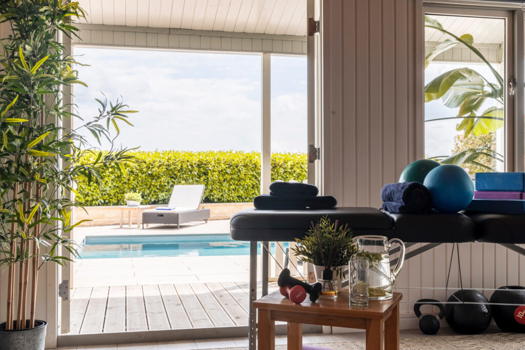 A massage table with folded towels, yoga balls, and fitness gear sits in a bright room overlooking a pool and lounge chair outside, with plants adding a relaxing, spa-like feel. David Pearson Travel