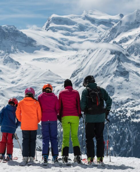 A group of five people in vibrant ski gear stand on a snowy mountain slope, indulging in the luxury travel experience amidst breathtaking snow-capped peaks under a partly cloudy sky. The vast landscape showcases mountains majestically stretching across the horizon. David Pearson Travel