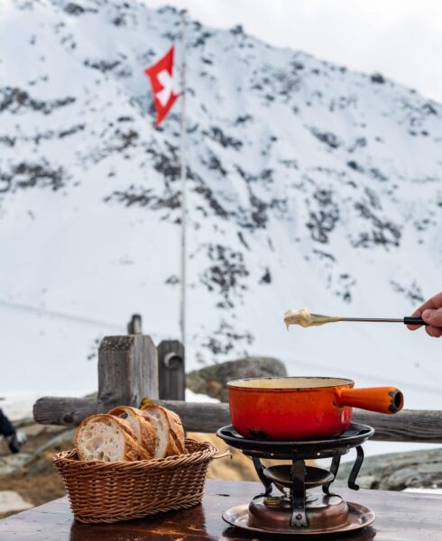 A pot of fondue sits elegantly on an outdoor table, framed by a breathtaking snowy mountain backdrop. A hand gracefully dips a fork with cheese above the pot, while a basket of sliced bread awaits beside it—a quintessential scene of Swiss luxury travel. David Pearson Travel