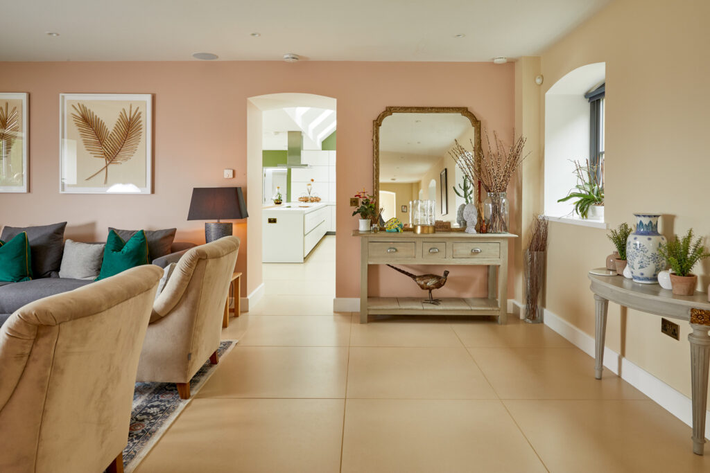 A stylish living room with beige armchairs, a gray sofa with green pillows, a wooden console with a mirror, decorative vases, and framed leaf art on a pale pink wall, leading to a modern white kitchen. David Pearson Travel