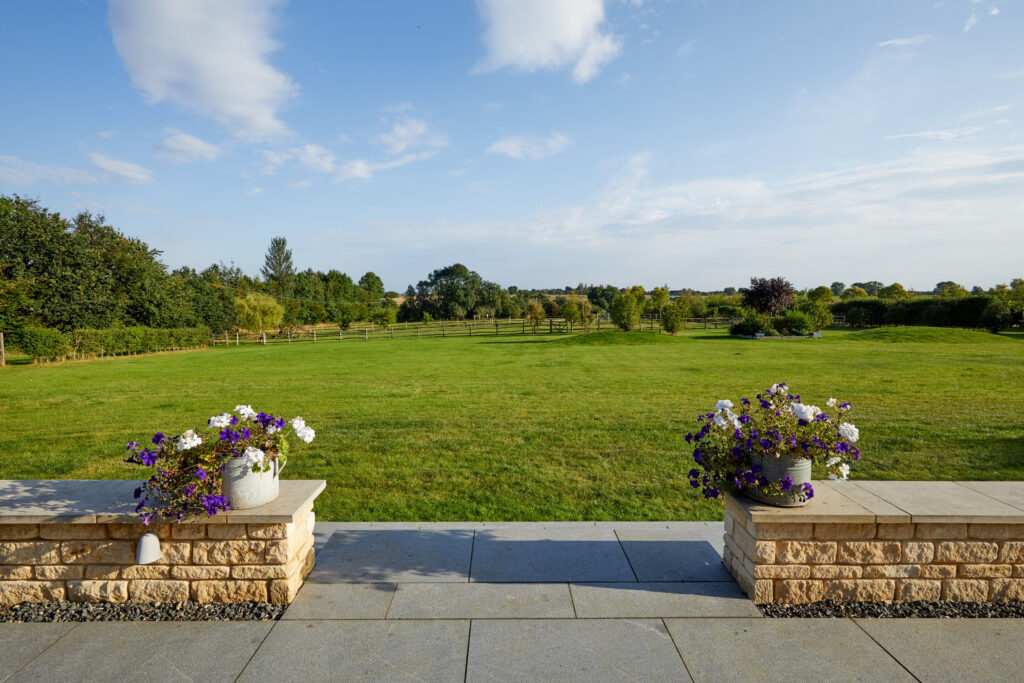 A lush green lawn stretches into the distance, bordered by trees under a blue sky. Two stone planters with purple and white flowers sit on a patio in the foreground. David Pearson Travel
