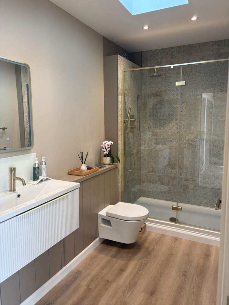 Modern bathroom with a floating vanity, wall-mounted toilet, walk-in glass shower with gold fixtures, light wood floor, and neutral walls. Decorative items, soap, and an orchid plant are on the counter. Skylight above. David Pearson Travel