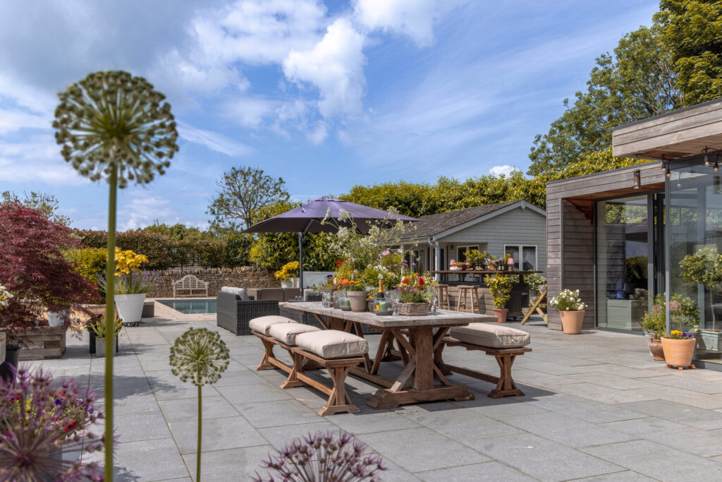 A sunny patio with wooden benches and a table set for outdoor dining, surrounded by potted plants, flowers, and a garden shed. An umbrella provides shade, and a pool is visible in the background. David Pearson Travel
