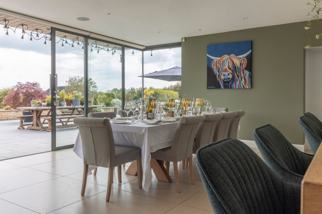 Modern dining room with a set dining table, grey upholstered chairs, glass doors opening to a patio with outdoor seating, and a colorful highland cow painting on a green wall. Lush greenery and flowers visible outside. David Pearson Travel