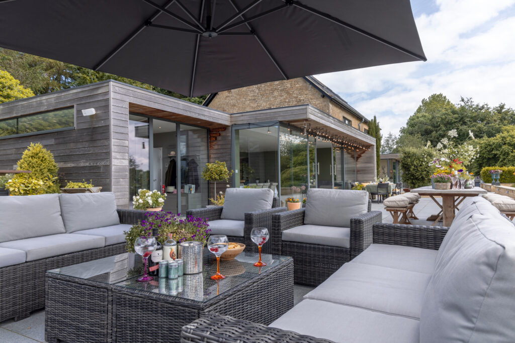 Modern outdoor patio with gray wicker furniture, glass-top table, and drinks under a large black umbrella. The patio is attached to a contemporary house with glass walls and surrounded by greenery. David Pearson Travel