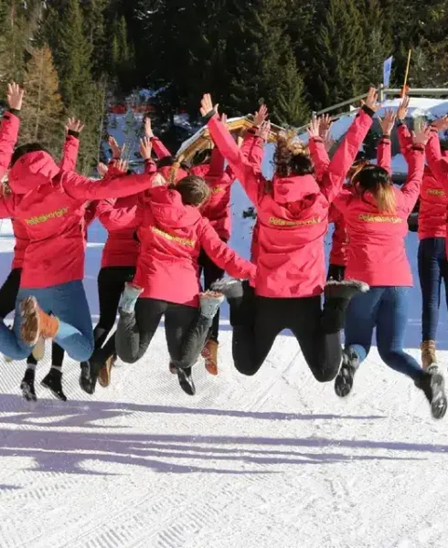 A group of people in matching red jackets jump in the air on a snowy slope in Verbier, facing away from the camera, with trees and blue sky in the background—a perfect moment for Verbier tourism. David Pearson Travel