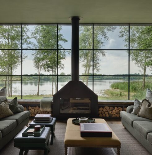 A modern living room with two gray sofas, a central fireplace, large windows revealing a lake and trees outside, and neatly stacked firewood under the windows. Pillows and books decorate the space. David Pearson Travel