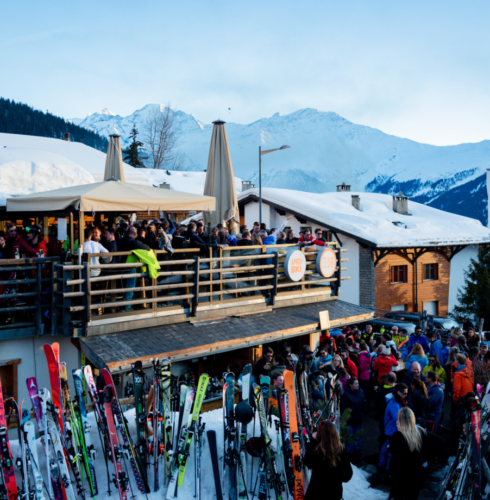 Crowds gather for après-ski at a lively outdoor alpine bar in Verbier, with snow-covered rooftops, colorful skis lined up outside, and snowy mountains in the background under a clear evening sky—perfect for Verbier travel enthusiasts. David Pearson Travel