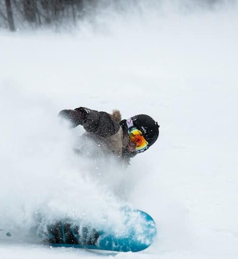 A snowboarder wearing goggles and winter gear kicks up a spray of snow while making a sharp turn on a snowy slope in Verbier, surrounded by a stunning winter landscape perfect for skiing and travel adventures. David Pearson Travel