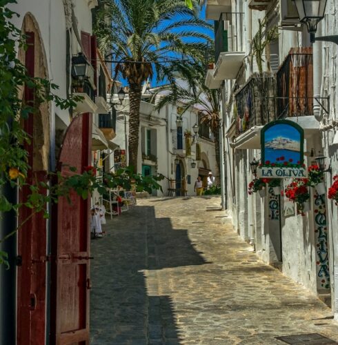 A narrow, sunlit cobblestone street winds uphill between whitewashed buildings with balconies, potted plants, and palm trees. A colorful La Oliva sign hints at the charm of Ibiza travel. The sky is clear and blue. David Pearson Travel