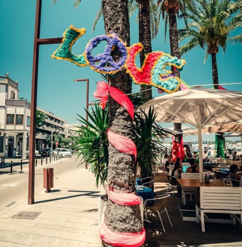 A palm tree on a sunny Ibiza street is decorated with colorful letters spelling LOVE and a peace sign, with pink fabric wrapped around the trunk. People sit at outdoor tables under umbrellas nearby. David Pearson Travel