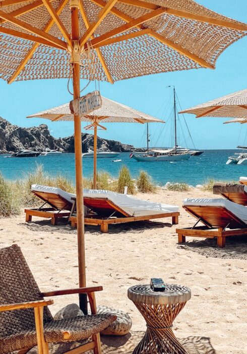 Beach scene with wooden lounge chairs and thatched umbrellas on sand, overlooking Ibiza’s blue sea with sailboats in the distance. Grassy dunes and a rocky hill are visible in the background under a clear sky. David Pearson Travel