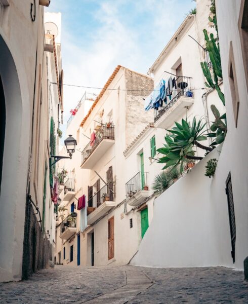 A narrow cobblestone street lined with whitewashed buildings, balconies displaying potted plants and colorful laundry, under a blue sky—an idyllic scene straight from an Ibiza guide for your next Ibiza travel adventure. David Pearson Travel