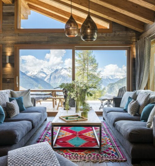 Chalet Mowgli David Pearson Travel A cozy living room perfect for your Home Page, with large windows revealing snowy mountains, two gray sofas, a colorful rug, and a wooden coffee table with flowers. Natural light highlights the wood beams and decorative pillows. David Pearson Travel