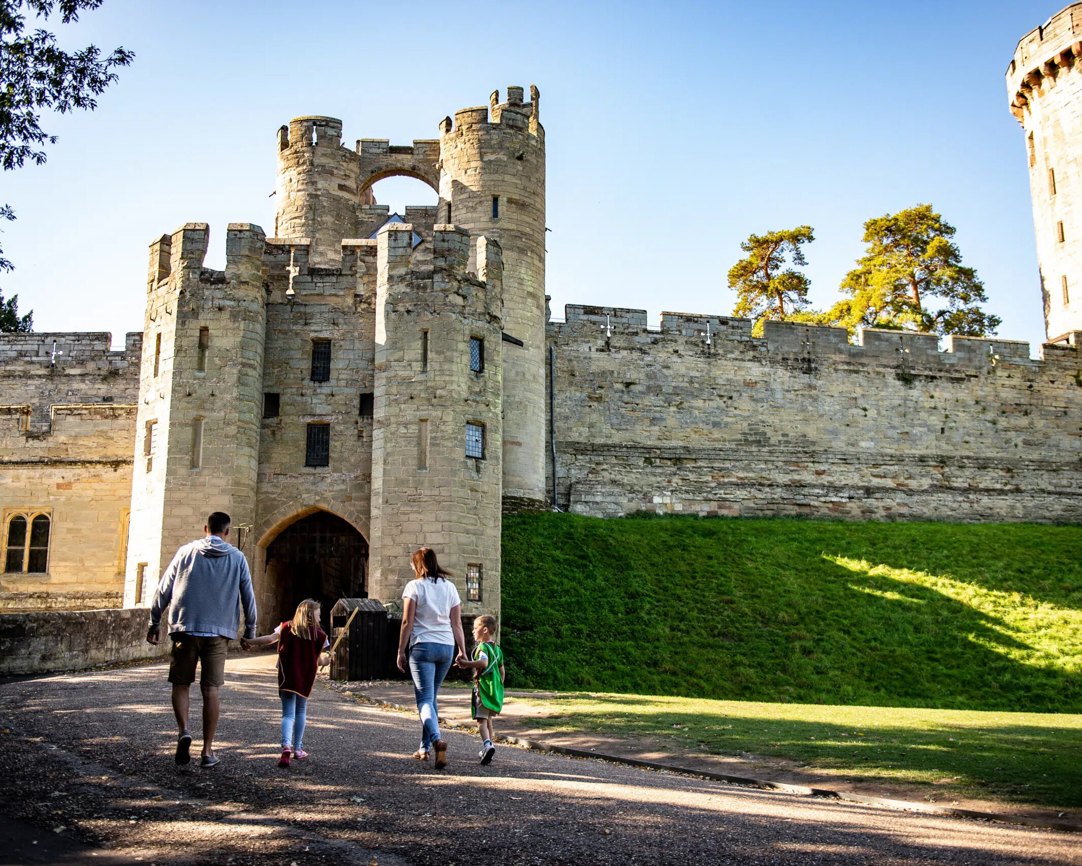 Cotswolds A family of four, two adults and two children, walk hand-in-hand toward the entrance of a large historic stone castle with towers and battlements on a sunny day, evoking the welcoming spirit of a Home Page. David Pearson Travel