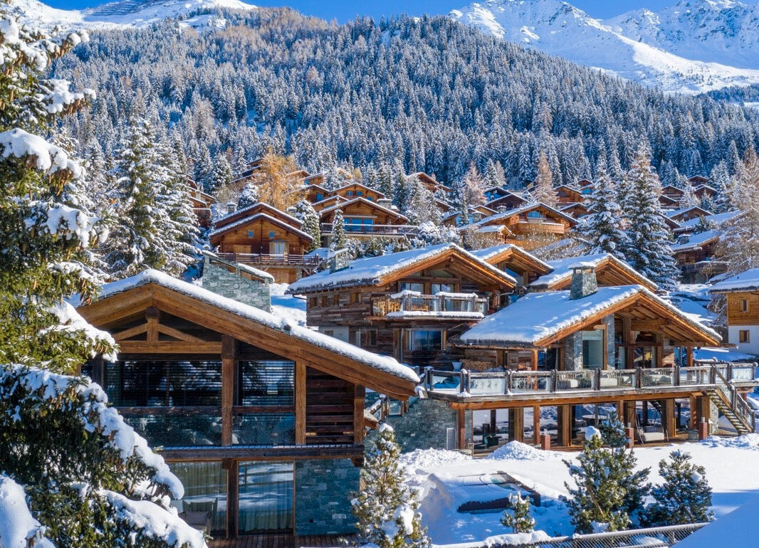 Verbier A cluster of wooden chalets with large windows sits in a snowy alpine village, perfect for your Home Page backdrop, surrounded by pine trees and mountains under a clear blue sky. Snow blankets rooftops and ground, creating a cozy wintery atmosphere. David Pearson Travel