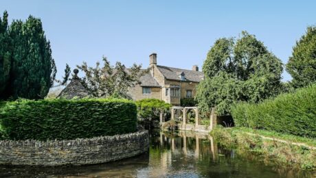 photo-1608221606535-15c75e4afb3c A stone cottage with a pitched roof sits beside a clear, shallow stream in The Cotswolds, surrounded by lush green trees and hedges under a bright blue sky. David Pearson Travel