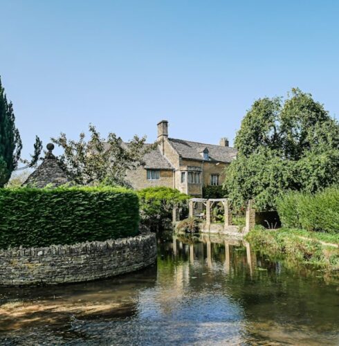 A stone cottage with a pitched roof sits beside a clear, shallow stream in The Cotswolds, surrounded by lush green trees and hedges under a bright blue sky. David Pearson Travel