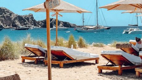 photo-1622890806166-111d7f6c7c97 Beach scene with wooden lounge chairs and thatched umbrellas on sand, overlooking Ibiza’s blue sea with sailboats in the distance. Grassy dunes and a rocky hill are visible in the background under a clear sky. David Pearson Travel
