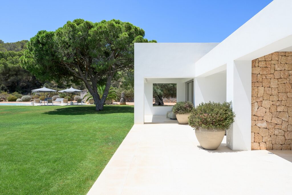 A modern white house with large windows and stone accents overlooks a green lawn with potted plants, a large tree, and outdoor seating with umbrellas in the background on a sunny day. David Pearson Travel