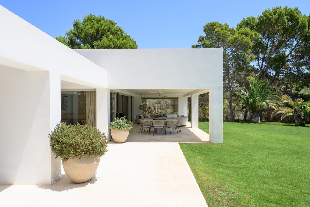 A modern white house with a covered patio, outdoor dining table and chairs, large potted plants, and a lawn bordered by trees under a clear blue sky. David Pearson Travel