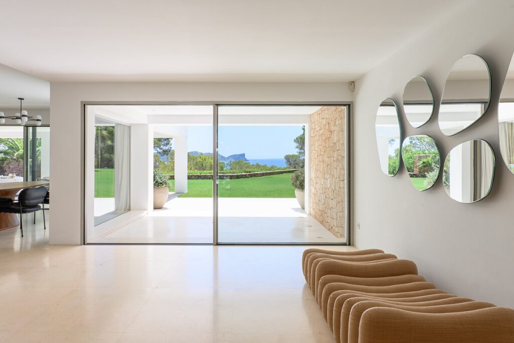 Modern, minimalist living space with tan wave-shaped seating, circular mirrors on a white wall, and large glass doors opening to a patio and lawn with a scenic sea and mountain view. David Pearson Travel