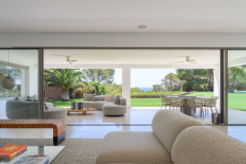 Modern living room with plush beige sofas opens to a spacious patio with round lounge chairs and outdoor dining set, overlooking a green lawn, palm trees, and a distant view of the sea. David Pearson Travel