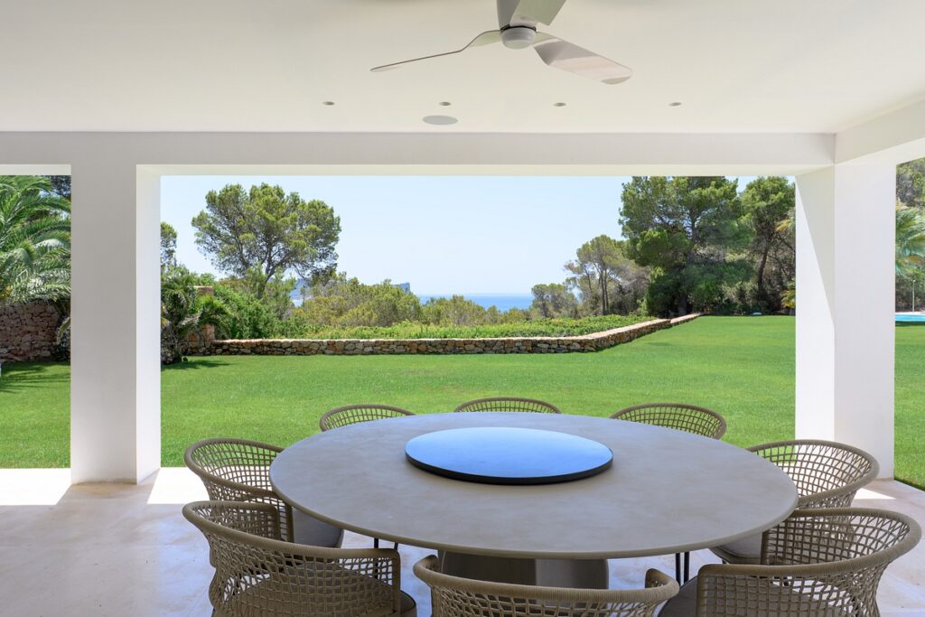 Covered patio with a round table and wicker chairs overlooking a large green lawn, stone wall, trees, and a distant view of the sea under a clear blue sky. David Pearson Travel
