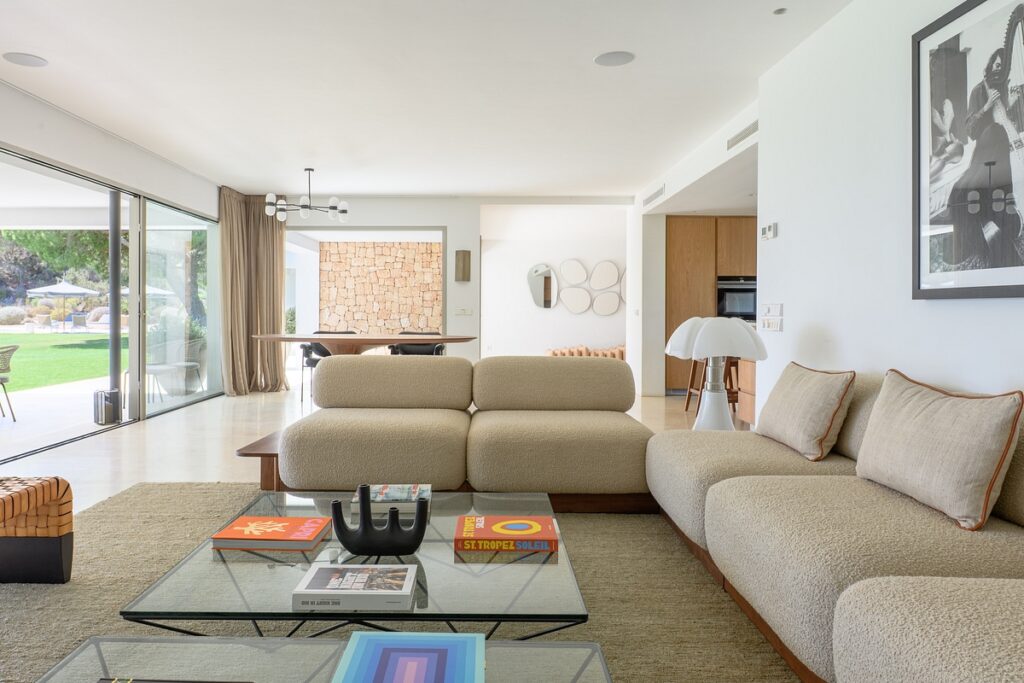 Modern living room with beige sofas, glass coffee table with books, large windows opening to a garden, and neutral decor. Dining area with a stone wall and contemporary light fixture is visible in the background. David Pearson Travel