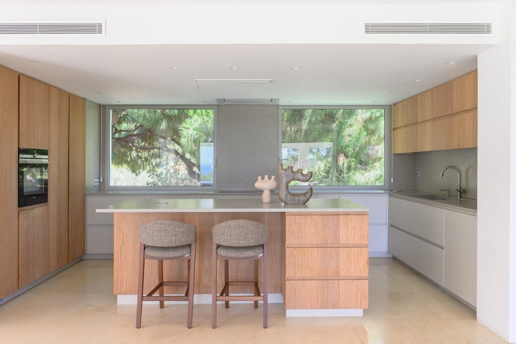 Modern kitchen with light wood cabinets, a central island with two cushioned stools, minimalist decor pieces on the island, and large windows showing green trees outside. The space is bright and airy. David Pearson Travel