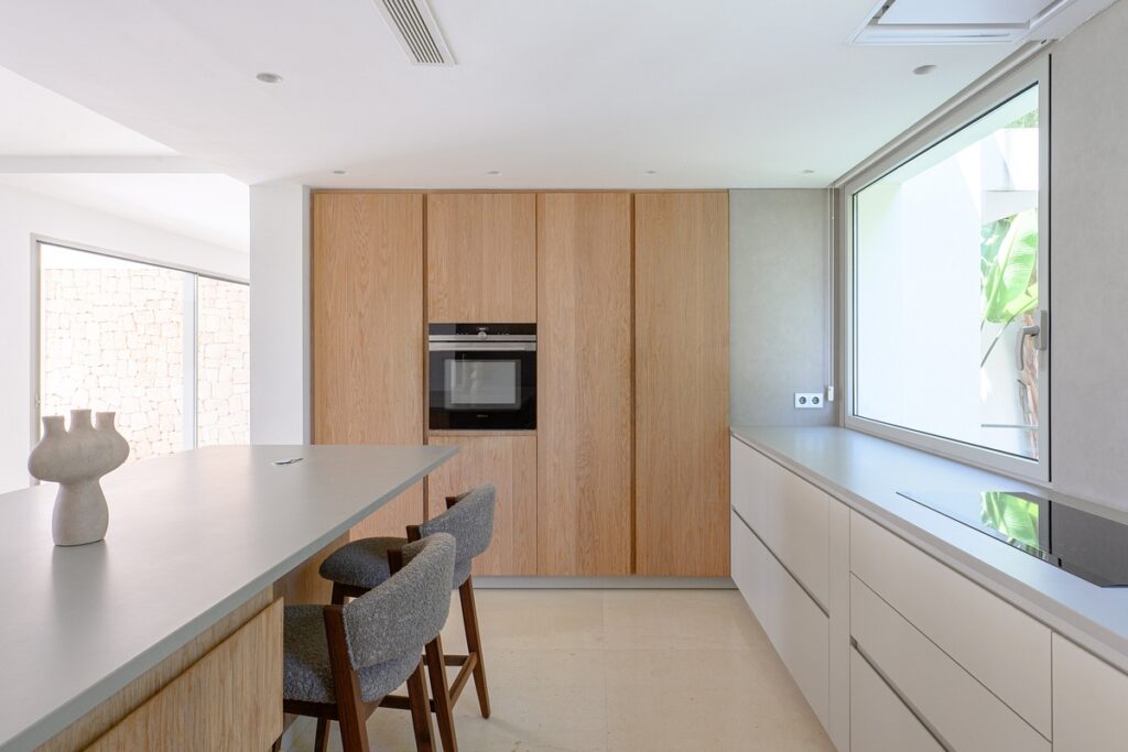 Minimalist kitchen with light wood cabinets, a built-in oven, a large window, a sleek countertop with two gray cushioned stools, and neutral-toned decor, creating a bright and modern atmosphere. David Pearson Travel