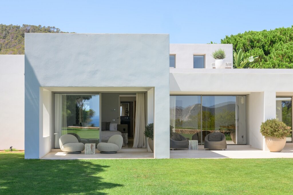A modern white villa with large glass doors, outdoor seating areas, and potted plants sits on a well-manicured lawn with green trees and hills visible in the background under a clear blue sky. David Pearson Travel