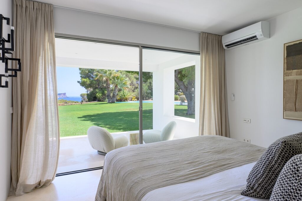 A modern bedroom with a large bed, neutral bedding, sliding glass doors, and light curtains opens to a patio with two round chairs and a view of a green lawn, trees, and distant water under a bright sky. David Pearson Travel