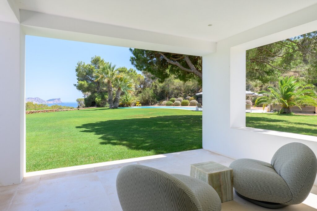 A modern covered patio with two cushioned chairs and a small table overlooks a lush green lawn, palm trees, and distant hills under a clear blue sky. David Pearson Travel