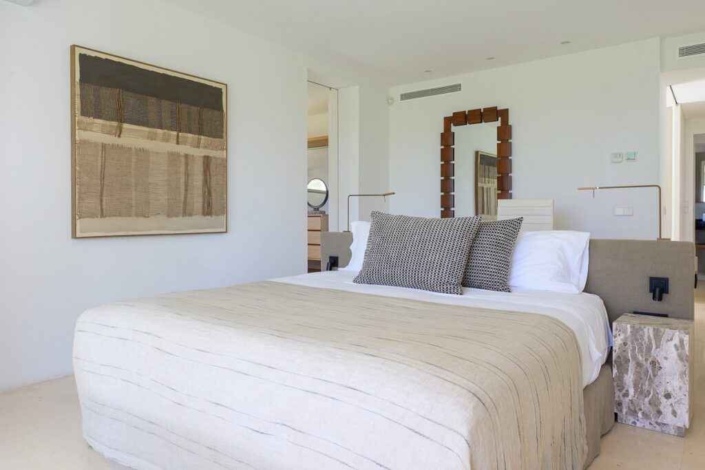 A modern, minimalistic bedroom with a neatly made bed, neutral bedding, patterned pillows, abstract wall art, a stone nightstand, and a large mirror with a geometric frame on a white wall. David Pearson Travel