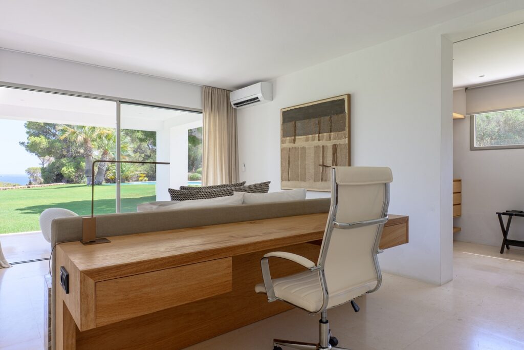 Modern living room with a wooden desk and white office chair in the foreground, a light sofa facing large glass doors that open to a green lawn and trees, and neutral decor with natural light filling the space. David Pearson Travel