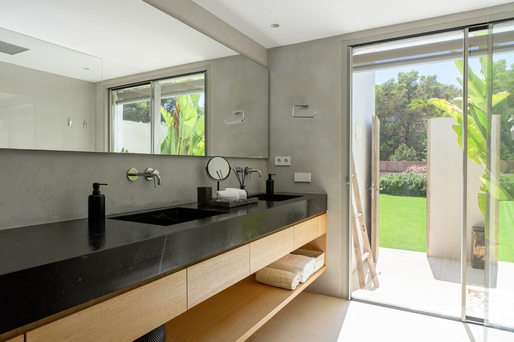 Modern bathroom with a black double sink countertop, wooden cabinets, large mirror, and glass door leading to a lush green garden with sunlight streaming in. Towels and soap dispensers are on the counter. David Pearson Travel