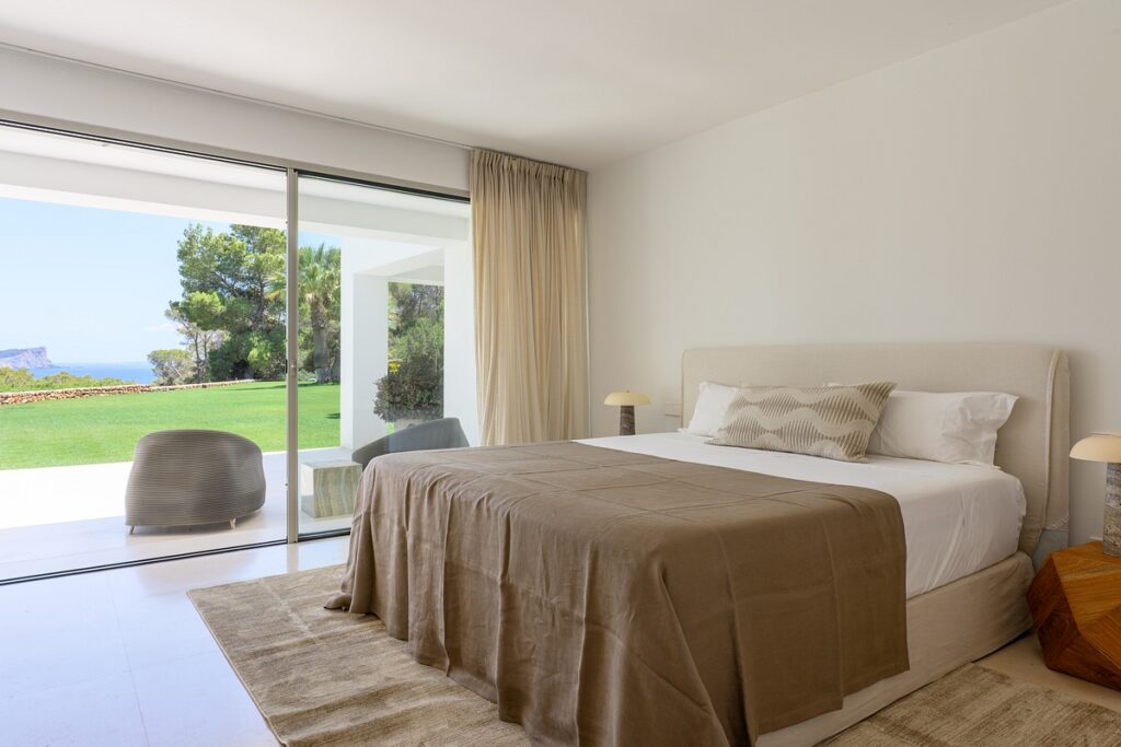A modern bedroom with a beige bedspread, neutral decor, large glass doors, and a view of a green lawn and trees outside. Light curtains, a rug, armchairs, and two bedside lamps complete the minimalist design. David Pearson Travel