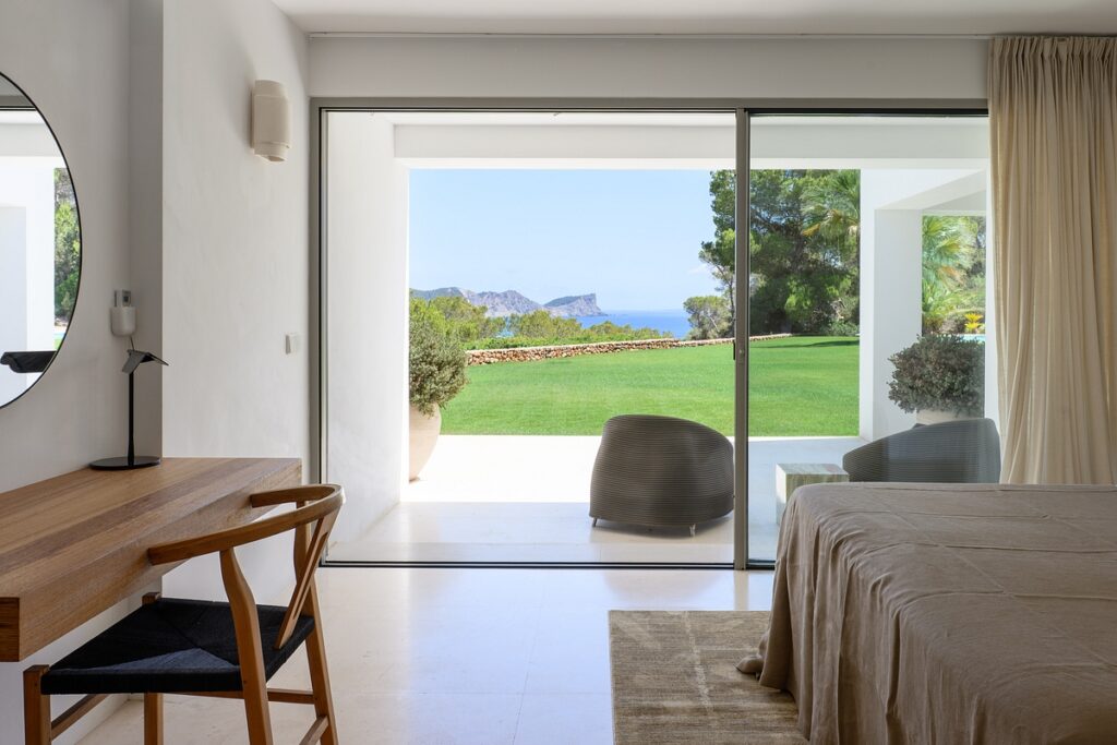 A modern bedroom with a wooden desk and chair, a bed, and large sliding glass doors opening to a green lawn, with trees and a view of the ocean and distant hills outside. David Pearson Travel