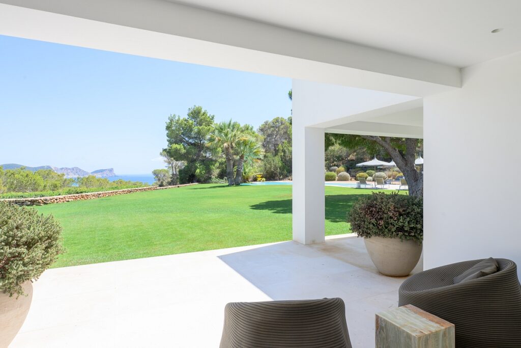 A covered patio with two chairs overlooks a lush green lawn, palm trees, and distant hills under a clear blue sky. The patio features large potted plants and has a view of the ocean in the background. David Pearson Travel