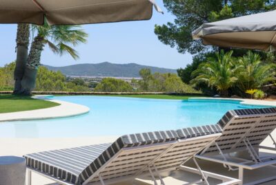 sacaleta-4 Three striped lounge chairs under umbrellas sit beside a curved swimming pool surrounded by palm trees and greenery, with distant mountains and a clear blue sky in the background. David Pearson Travel