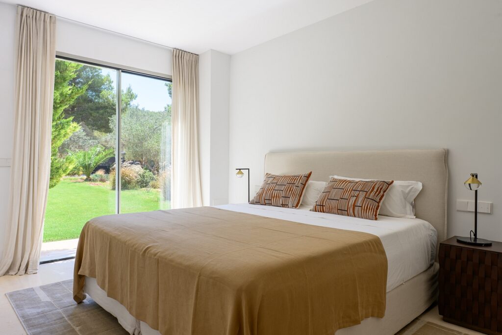 A bright bedroom with a large bed covered in a beige blanket and patterned pillows. The room has white walls, light curtains, and a sliding glass door that opens to a green garden outside. David Pearson Travel