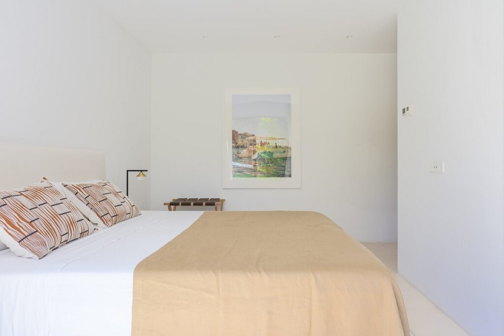 Minimalist bedroom with white walls, a bed with a beige blanket and patterned pillows, a small wall lamp, a bench, and a framed artwork hanging above the bench. The room is bright and uncluttered. David Pearson Travel