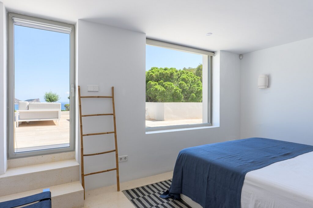 Minimalist bedroom with white walls, a bed with a blue blanket, a striped rug, a wooden ladder, and large windows showing a patio with outdoor seating and greenery under a clear blue sky. David Pearson Travel